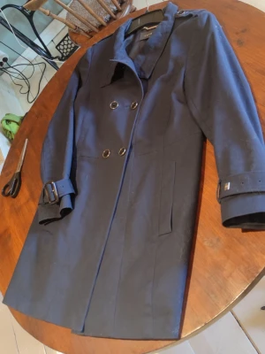 Mörkblå trenchcoat med bältdetaljer - Stilren mörkblå trenchcoat från Vila med dubbelknäppning, bälten vid ärmslut och midja samt klassisk krage. Jackan har raka fickor och en snygg slits bak. Perfekt för dig som vill ha en tidlös och trendig look. Storlek passar även den som har storlek M