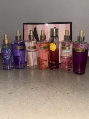 Body mists från Victoria's Secret - Säljer ett set med sju olika body mists från Victoria's Secret i färger som lila, rosa och röd. Flaskorna är genomskinliga med guldfärgade spraymunstycken och har olika doftnamn på etiketten. Perfekt för dig som gillar fräscha och blommiga dofter.
