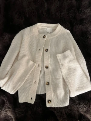 Krämvit kofta från H&M - Mysig krämvit kofta från H&M med ribbstickad struktur och stora bruna knappar framtill. Koftan har rund halsringning och långa ärmar, knappt andvänd!
