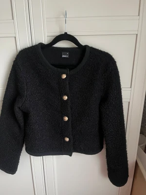 Svart bouclékofta från Gina Tricot - Stilren svart bouclékofta från Gina Tricot med rund halsringning och fyra dekorativa guldfärgade knappar framtill. Koftan har lång ärm och en croppad passform. Perfekt att slänga över en topp för en trendig look.