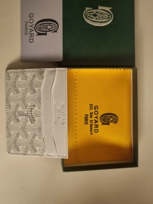 Vit kortfodral från G4yard - Säljer ett stilrent vitt kortfodral från G4yard med det klassiska mönstret i grått och svart. Kortfodralet har flera fack för kort och är tillverkat i exklusivt läder. Kommer med originalask och gul putsduk. Perfekt för dig som vill ha något lyxigt och tidlöst.