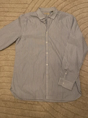 Randig skjorta från Burberry London - Klassisk skjorta från Burberry London i vitt och blått med smala vertikala ränder. Skjortan har lång ärm, knappar framtill och en tidlös krage. Tillverkad i mjuk bomull för en bekväm känsla och stilren look.