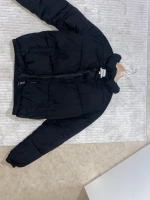 Svart pufferjacka från Kappahl  - Säljer en svart pufferjacka från Kappahl med hög krage och dragkedja framtill. Jackan har två sidofickor och en reflekterande detalj på ryggen. Perfekt för kalla vinterdagar och riktigt skön att bära tack vare det vadderade materialet. Den är använd bara 2 gånger, alltså helt ny! Meddela vid frågor :)