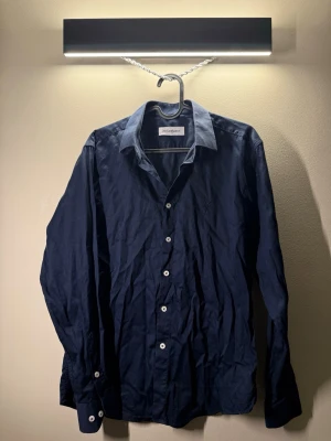 Mörkblå skjorta från Yves Saint Laurent - Snygg mörkblå skjorta från Yves Saint Laurent med klassisk krage och vita knappar. Skjortan har lång ärm och diskret YSL-logga broderad på bröstet. Perfekt för dig som gillar stilrena och exklusiva plagg.