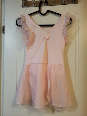 Dansdräkt ca strl 140 - Söt ljusrosa kortklänning med volangärmar och v-ringning. Klänningen har en liten rosett framtill och är gjord i ett tunt, lätt material med ett lager av transparent tyg över kjolen och ärmarna. Perfekt för dans eller balett. Ca storlek 140.