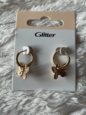 Guldiga örhängen med fjärilar Glitter - Snygga guldiga hoops från Glitter med detaljerade fjärilsberlocker. Örhängena är i metall och har en rund form med hängande fjärilar som ger en trendig och söt look. Perfekta för att lyfta vilken outfit som helst. 🌸