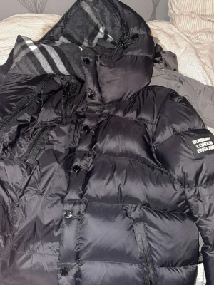 Svart pufferjacka från Burberry - Säljer en svart pufferjacka från Burberry med huva och dragkedjor framtill. Jackan har en patch med texten 'Burberry London England' på ärmen och stora, vadderade paneler för extra värme. Perfekt för kalla vinterdagar. 