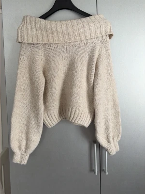 Beige off shoulder stickad tröja - Säljer en beige stickad tröja från Gina Tricot med bred off shoulder-krage och ballongärmar. Tröjan har ribbstickade muddar och är gjord i mjukt garn, perfekt för kalla dagar. Enkel att styla till jeans eller kjol.