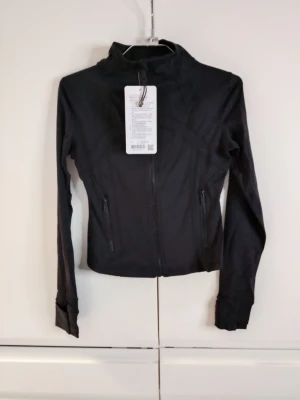 Svart träningsjacka från Lululemon Size 4 - Snygg svart träningsjacka från Lululemon med hög krage och dragkedja framtill. Jackan har två fickor med dragkedja och är tillverkad i ett stretchigt funktionsmaterial som passar perfekt till träning eller löpning.