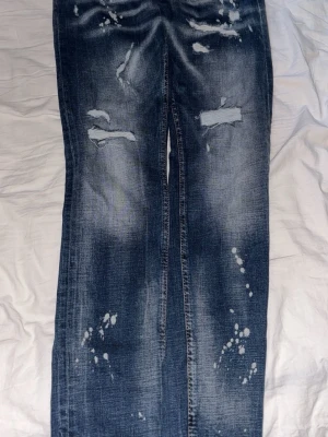 Dsquared2 slitna blå jeans - Säljer ett par blå jeans från Dsquared2 med slitna detaljer, hål och blekta partier. Jeansen har raka ben och klassisk femficksmodell. Snygga kontrastsömmar och coolt tvättad denim ger en trendig look.