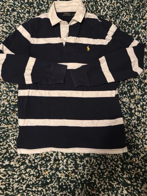 Randig långärmad skjorta Polo Ralph Lauren - Mörkblå och vit randig långärmad skjorta från Polo Ralph Lauren med klassisk krage och knappar framtill. Ikoniska gula loggan broderad på bröstet. Tillverkad i mjuk bomull, perfekt för en avslappnad men ändå stilren look.