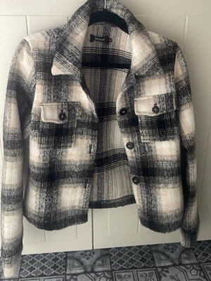 Rutig skjorta från ONLY i svart och vitt - Säljer en snygg rutig skjorta från ONLY i svart, vitt och grått. Skjortan har klassisk krage, knappar framtill och två bröstfickor med lock. Perfekt för lager på lager och ger en cool, avslappnad look.