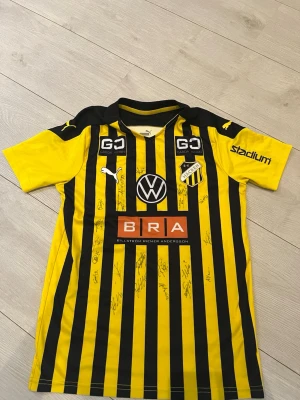 BK Häcken matchtröja Puma gul/svart - Säljer en BK Häcken matchtröja med alla autografer från Puma i gul och svart med vertikala ränder. Tröjan har flera sponsorloggor, klubbmärke på bröstet och tryckta autografer. Tillverkad i funktionsmaterial och har korta ärmar samt rund hals.