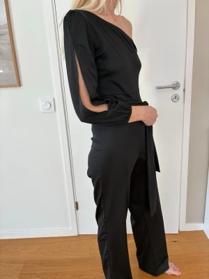 Svart jumpsuit one shoulder stl S - Svart jumpsuit med en axel och lång ärm med slits. Jumpsuiten har ett knytband i midjan. Polyester. Använd en gång, är i mkt bra skick. Inga defekter. Stl S.