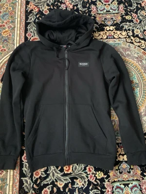 Svart zip hoodie från McKenzie - Säljer en svart hoodie från McKenzie med dragkedja framtill och stor huva. Hoodien har en liten patch med McKenzie-loggan på bröstet och två rymliga fickor framtill. Tillverkad i mjukt bomullsmaterial som är skönt att ha på sig.