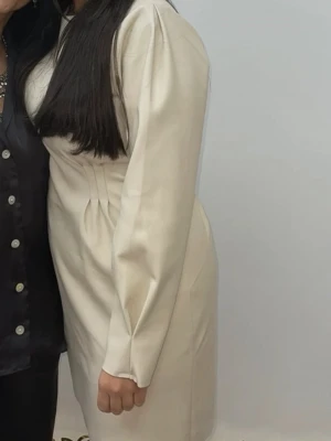 Krämvit långärmad kortklänning - Säljer en stilren krämvit kortklänning med långa puffärmar och markerad midja. Klänningen har en enkel, elegant design utan synliga detaljer och är perfekt för dig som gillar minimalistisk stil. Den har en mjuk foder inne så ingen läder känsla när man bär den. Från Nelly 