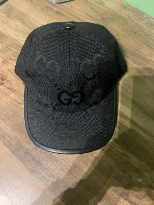 Svart keps från Gucci med GG-mönster - Snygg svart keps från Gucci med det klassiska GG-mönstret ton-i-ton över hela kepsen. Justerbar rem bak i läder och böjd skärm. Tillverkad i slitstarkt tyg som ger en exklusiv känsla. Perfekt för dig som vill ha en stilren accessoar med lyxig touch.