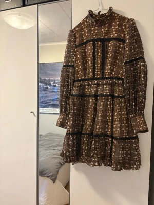By Malina klänning, slutsåld - By Malina klänning, slutsåld. Kort klänning i brunt och beige med små vita blommor, volangdetaljer och svarta band. Hög krage och långa ärmar med lätt puff. Klänningen har en luftig passform och är tillverkad i ett lätt material, perfekt för vår och höst.