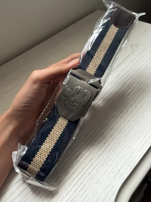 Randigt marinblått tygbälte med metallspänne - Snyggt tygbälte i marinblått och beige med randigt mönster och ett coolt metallspänne med graverad logga. Perfekt för att ge din outfit en sportig touch. Justerbar längd och slitstarkt material.