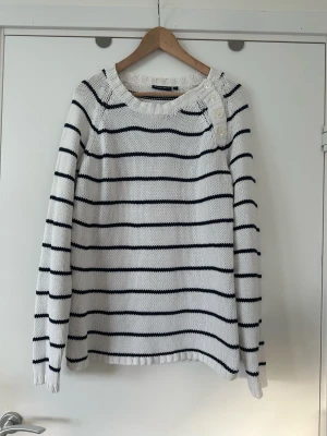 Vit stickad tröja med marinblå ränder, Twilfit, L - Klassisk vit stickad tröja från TWILFIT med marinblå horisontella ränder i storlek L. Tröjan har långa ärmar, ribbstickad halsringning och dekorativa knappar på ena axeln. Perfekt för en stilren och tidlös look. Mycket fint skick. 