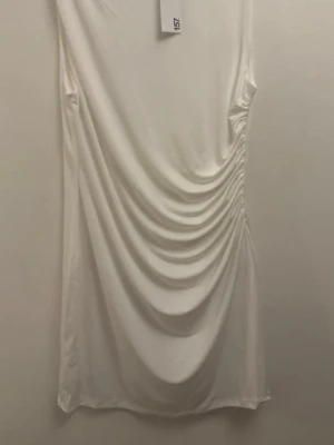 Vit kortklänning med drapering - Snygg vit kortklänning med draperad effekt på sidan som ger en cool och trendig look. Klänningen har en enkel, ärmlös design och är tillverkad i ett mjukt, stretchigt material som sitter skönt på kroppen. Perfekt för dig som gillar minimalistisk stil med en twist.