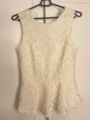 Vit spetspeplumtop med öppen rygg - Snygg vit peplumtop i spets med ärmlös design och rund halsringning. Har ett elegant öppet parti i ryggen och dragkedja baktill. Materialet är spets med blommönster och insida i slätt tyg. 
