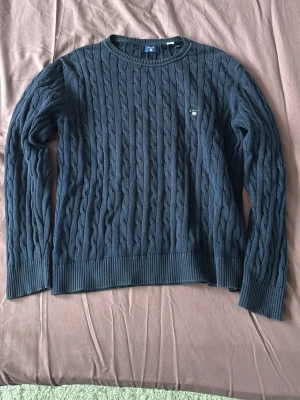 Mörkblå kabelstickad tröja Gant - Säljer en mörkblå kabelstickad tröja från Gant med rund hals och diskret logotyp broderad på bröstet. Tröjan har långa ärmar och ribbade muddar vid ärmslut och nederkant. Perfekt för dig som gillar klassisk och stilren design.