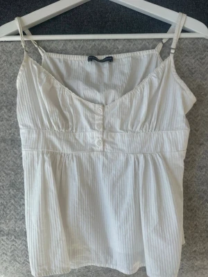 Vit randig linnetopp Brandy Melville - Söt vit linnetopp från Brandy Melville med tunna axelband och subtila vertikala ränder. Toppen har knappar framtill och en lätt rynkad byst för en avslappnad och luftig känsla. Perfekt för varma dagar och enkel att styla.