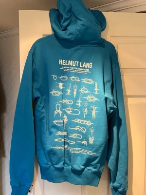 Helmut Lang hoodie  - Säljer en blå hoodie från Helmut Lang med stor huva och magficka. På bröstet finns ett vitt tryck med texten 'Helmut Lang Lifeguard' och ett kors, medan ryggen har ett stort grafiskt tryck med olika illustrationer och text. Tillverkad i mjukt bomullsmaterial och har ribbade muddar.