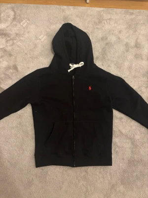 Ralph Lauren Zip - Svart hoodie med dragkedja från Polo Ralph Lauren. Klassisk design med vit dragsko i huvan och röd broderad logga på bröstet. Mjuk bomullskänsla och känguruficka framtill. Perfekt för en avslappnad och stilren look.