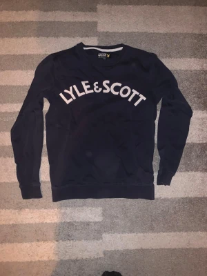 Mörkblå sweatshirt från Lyle & Scott - Mörkblå sweatshirt från Lyle & Scott med stor vit logotyp broderad över bröstet. Tröjan har rund halsringning, långa ärmar och ribbade muddar vid ärmslut och nederkant. Perfekt för en avslappnad och stilren look. DM för tydligare bild på bröstet, logotypen är broderad på ett speciellt sätt! 