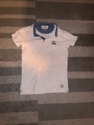 Vit pikétröja från Hampton Republic - Snygg vit pikétröja från Hampton Republic med blå detaljer på kragen och insidan av knäppningen. Tröjan har korta ärmar och en liten logga på bröstet. Perfekt för en clean och klassisk stil. Materialet känns mjukt och bekvämt.