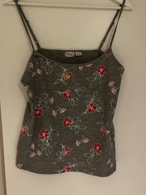 Grå blommig linnetopp från Esmara - Snygg grå linnetopp från Esmara med tunna axelband och blommigt mönster i rött, vitt och rosa. Toppen har små knappar framtill och är perfekt för varma dagar. Bander har gått sönder liter där framme, det finns en bild.