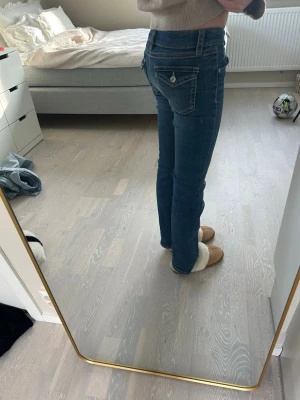 Mörkblå bootcut jeans med fickdetaljer - Säljer ett par mörkblå bootcut jeans med snygga kontrastsömmar och dekorativa fickor bak med knappdetalj. Jeansen har låg midja och klassisk femficksmodell. Perfekta för dig som gillar en tidlös och avslappnad stil. 