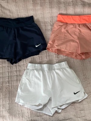 Nike sportshorts i tre färger - Säljer tre par sportiga shorts från Nike i färgerna marinblå, korall och vit. Alla har elastisk midja och är tillverkade i ett lätt, snabbtorkande material. De är tennisshorts men funkar även för annan träning. Klassisk Nike-logga på benet för en sportig look. De i marinblå och de korall är samma modell medan de vita har en liten annorlunda midja.