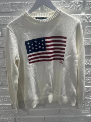 Vit stickad tröja Ralph Lauren USA-flagga - Vit stickad tröja från Ralph Lauren med stor amerikansk flagga i rött, blått och vitt på bröstet samt RL-broderi. Tröjan har rund halsringning och ribbade muddar vid ärmslut och nederkant. Perfekt för dig som gillar klassisk preppy stil.