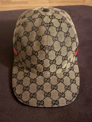 Navy blue gucci keps med mönster - Säljer en beige keps från Gucci med det klassiska GG-monogrammet över hela kepsen. Kepsen har en rak skärm och röda samt gröna detaljer på sidan. Materialet är canvas med läderdetalj på toppen. Perfekt för dig som gillar exklusiv streetwear.