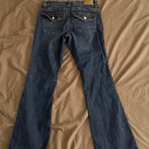 Blå bootcut jeans  - Säljer ett par klassiska blå bootcut jeans från Gina Tricot med två bakfickor som har lock och knappar. Jeansen är använda men i ett bra skick.