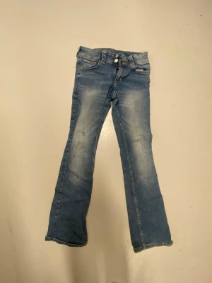 Blå bootcut jeans med dubbla knappar - Säljer ett par blå bootcut jeans med snygg slitning och dubbla knappar i midjan. Jeansen har klassisk femficksmodell och dragkedja. Perfekta för dig som gillar en retro och avslappnad stil.