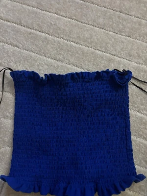 Blå bandeau tubtopp med volang - Säljer en blå bandeau tubtopp med smockad struktur och volangkant upptill och nedtill. Enkel och trendig modell som passar till mycket.