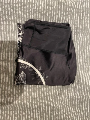 Svarta leggings från Anarchy Apparel - Snygga svarta leggings från Anarchy Apparel med vit blommig print och stor logga längs benet. Tillverkade i stretchigt material som sitter skönt och följer kroppen. Perfekta för träning. Unik design med detaljerade blommor och kontrasterande sömmar.