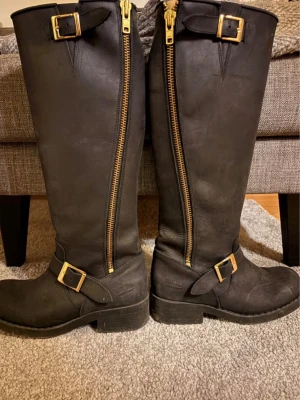 Svarta höga läderstövlar med dragkedja - Säljer ett par svarta höga stövlar från Johnny bulls i robust läder. Stövlarna har grov sula, dekorativa spännen i guld och dragkedja längs sidan. Perfekta för en edgy look och funkar till både jeans och kjol.