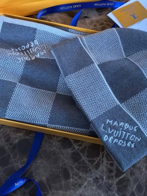 Grå rutig halsduk Louis Vuitton - Säljer en exklusiv grå halsduk från Louis Vuitton med klassiskt rutmönster och broderad text 'Marque L. Vuitton Depose'. Halsduken är mjuk och stilren, perfekt för att lyfta din outfit. Materialet är troligtvis ull eller en ullblandning.