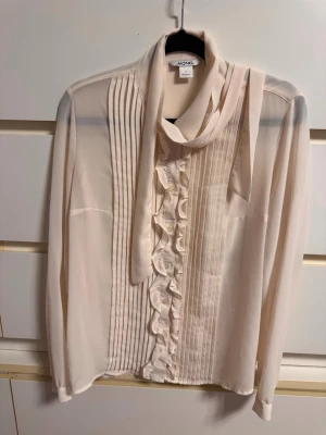 Krämvit blus med knytband MONKI - Elegant krämvit blus från MONKI med långa ärmar, knytband i halsen och dekorativa volanger samt veck framtill. Tillverkad i ett tunt, lätt material som ger en luftig känsla. Perfekt för en chic och stilren look.