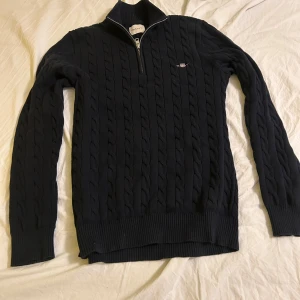 Svart kabelstickad tröja med halvzip - Säljer en svart kabelstickad tröja från Gant med halvzip och hög krage. Tröjan har ett diskret broderat Gant-logo på bröstet och ribbade muddar vid ärmslut och nederkant. Perfekt för dig som gillar klassisk och stilren design.