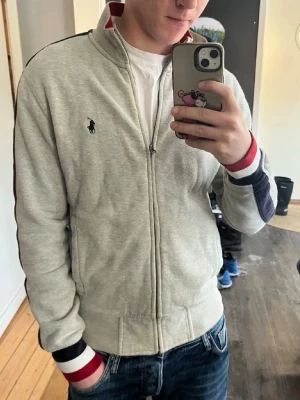 Grå hoodie från Ralph Lauren - Snygg grå hoodie från Ralph Lauren med dragkedja och hög krage. Har klassisk Polo-logga på bröstet och ribbade muddar med röd, vit och marinblå detalj. Perfekt för en chill och stilren look. Tillverkad i mjukt bomullsmaterial.