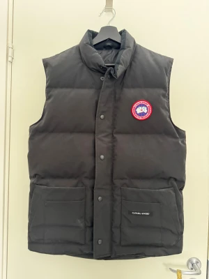 Svart dunväst från Canada Goose - Säljer en svart dunväst från Canada Goose med hög krage, två stora framfickor och klassisk logotyp på bröstet. Västen har tryckknappar framtill och är riktigt puffig och varm, perfekt för kyliga dagar. Materialet är slitstarkt och fyllningen är dun. Är det några frågor så tveka inte på att skriva! Kvitto finns ej tyvärr!