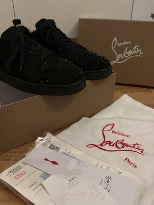Christian Louboutin svarta spikes sneakers - Svarta sneakers från Christian Louboutin med ikoniska röda sulor och svarta nitar över tån. Skorna har lågt skaft, snörning och är tillverkade i mocka och läder. Perfekta för dig som vill sticka ut med en edgy och lyxig stil. Pris kan diskuteras vid snabb affär