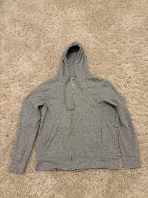 Grå zip hoodie från Uniqlo - Snygg  hoodie från Uniqlo med dragkedja framtill och justerbar huva. Tillverkad i mjukt bomullsmaterial som känns skönt mot huden. Perfekt för chill dagar eller när du vill ha en enkel och clean look.