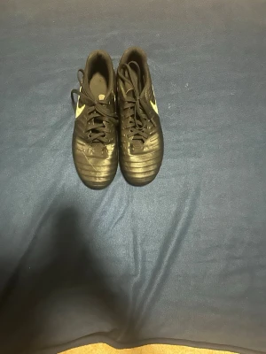 Svarta Nike fotbollsskor med dobbar - Säljer ett par svarta fotbollsskor från Nike med guldfärgade detaljer och klassisk swoosh på sidan. Skorna har snörning och fasta dobbar för spel på gräs. Tillverkade i syntetmaterial med en stilren och sportig design som passar perfekt till match eller träning.